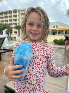 Young girl holding a blue icy in a bathing suit with flowers smiling with blonde straight hair that is wet with a water park behind her.