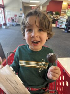 Pediatric aged boy smiling with brown curly hair holding a chocolate cake pop in a grocery cart at the store.