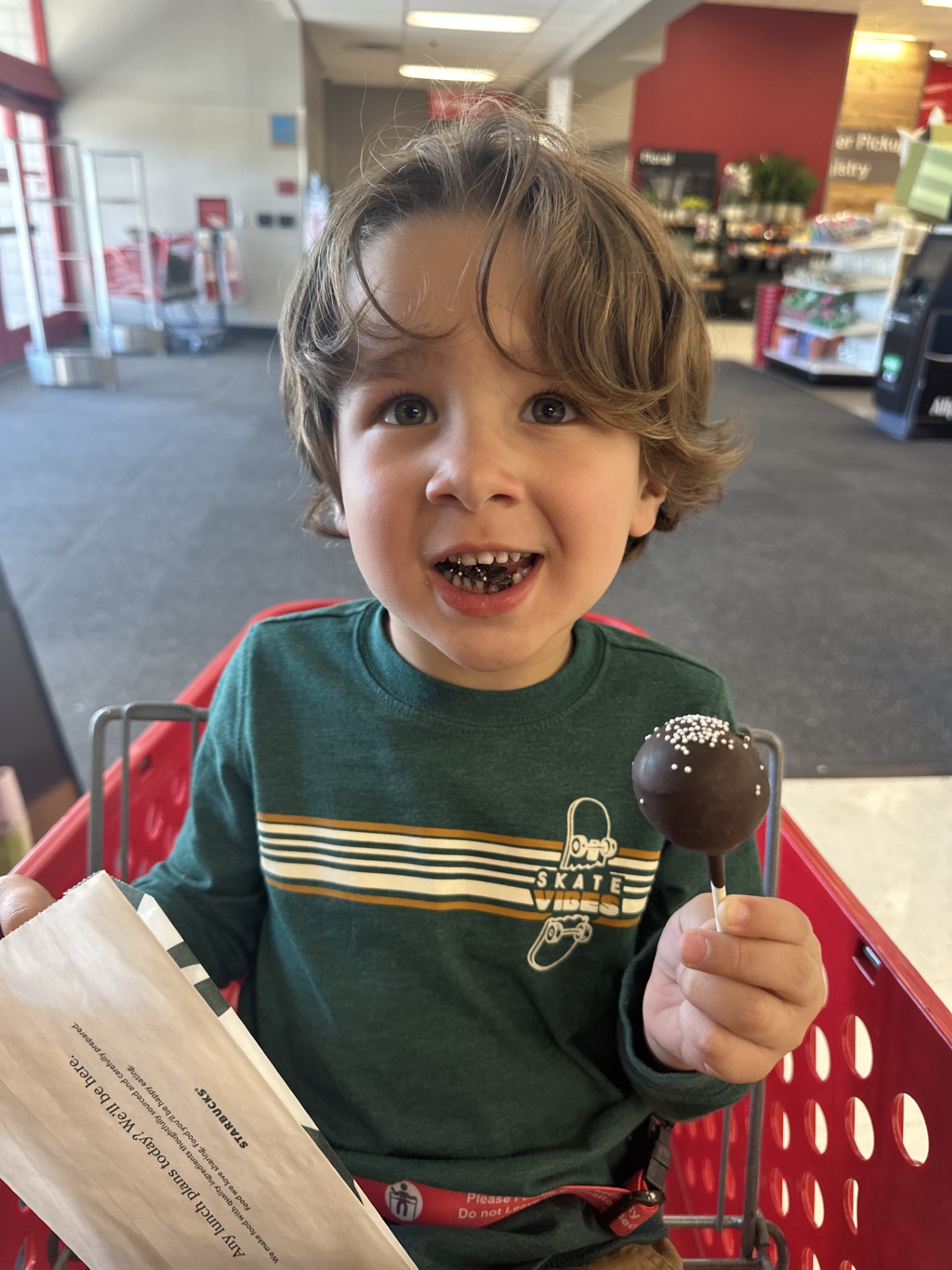 Pediatric aged boy smiling with brown curly hair holding a chocolate cake pop in a grocery cart at the store.