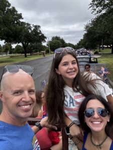 Winston Sheen, pediatrician, smiling, wearing sunglasses on his forehead next to a pediatric aged girl who is smiling with sunglasses on top of her brown hair in a USA flagged shirt, next to a woman with brown hair, smiling, with sunglasses on.