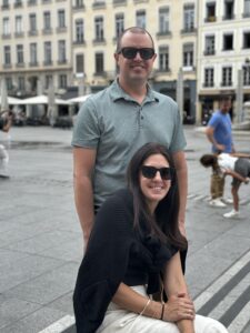 Winston Sheen, First call pediatrics pediatrician, is smiling with sunglasses on, in a grey shirt, behind a woman smiling, wearing sunglasses, in a black jacket, sitting on a bench, in front of buildings.