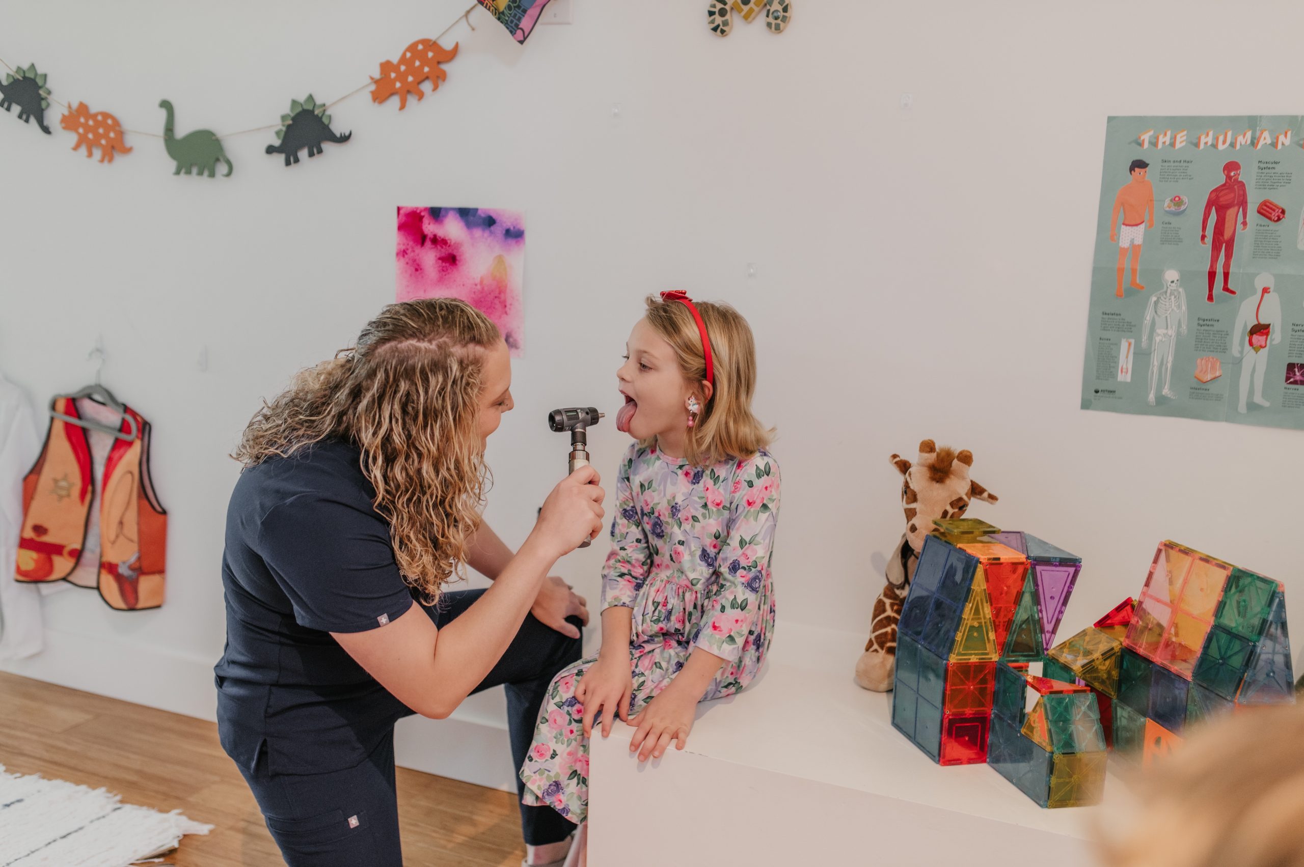Female NNP is in First Call Pediatric scrubs, kneeling, holding a laryngoscope and is preforming physical exam on 6-year-old girl whose tongue is out, in child’s room, in-home in San Marco neighborhood in Jacksonville, Fl.