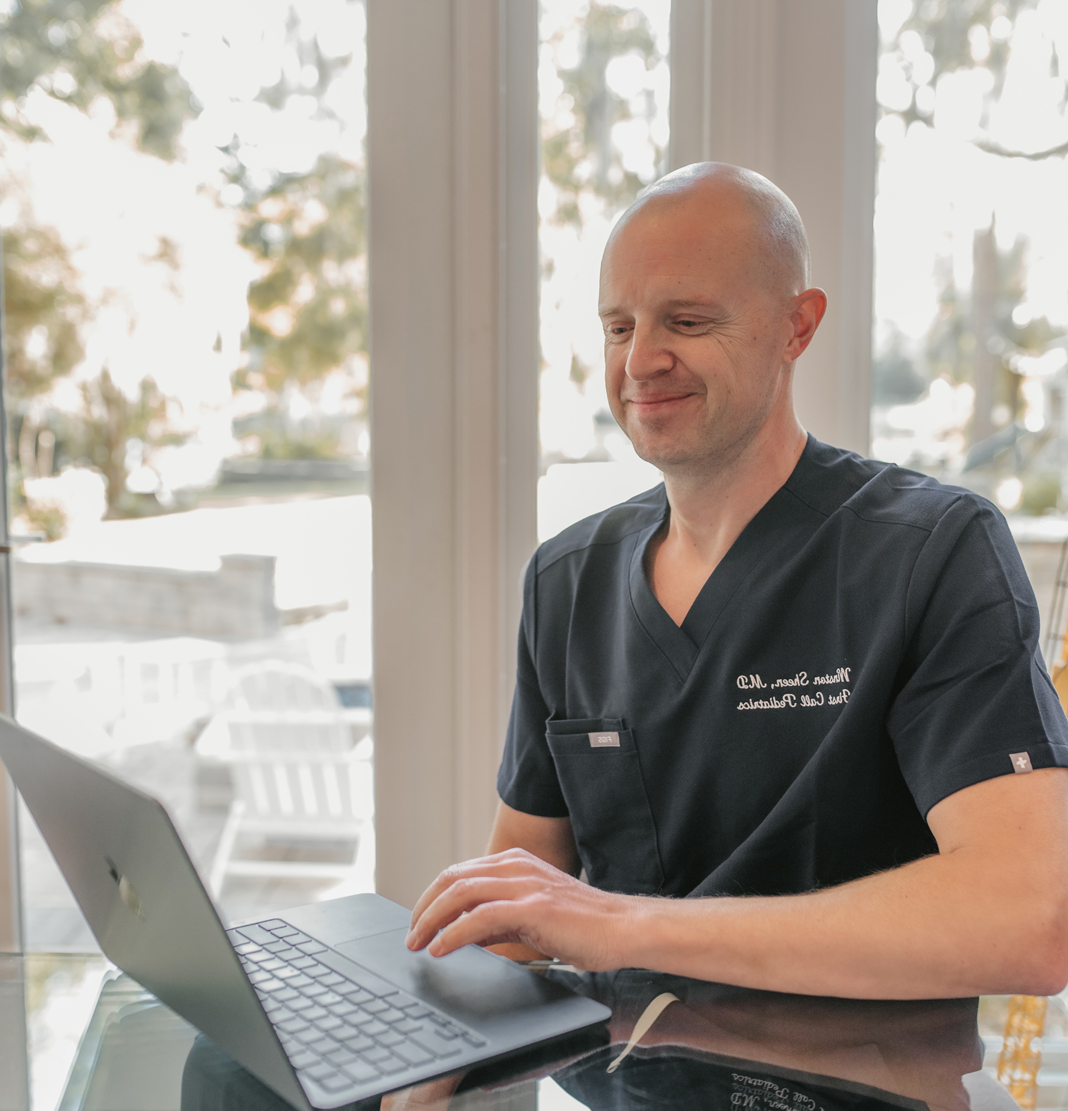 Male Pediatric Doctor is in First Call Pediatric scrubs, smiling, while using laptop in-home in San Marco neighborhood in Jacksonville, Fl.