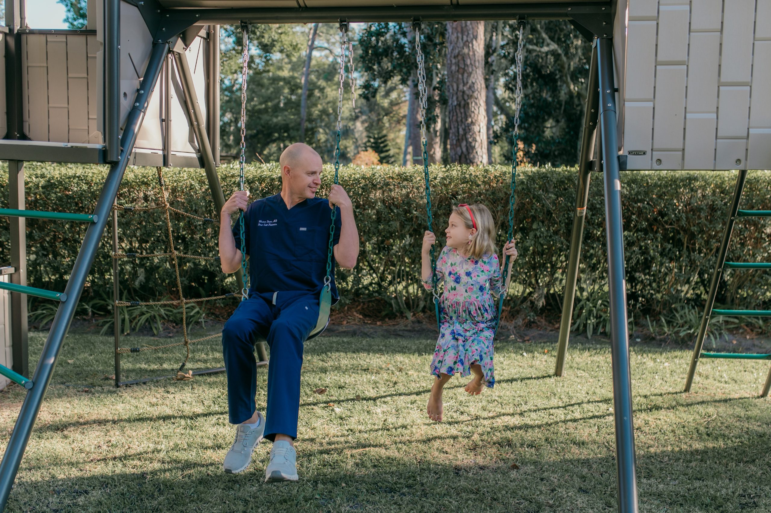 Male pediatric doctor, in First Call Pediatrics scrubs, smiling, sitting next to and looking at 6-year-old girl, smiling, outside at home in San Marco neighborhood in Jacksonville, Fl.