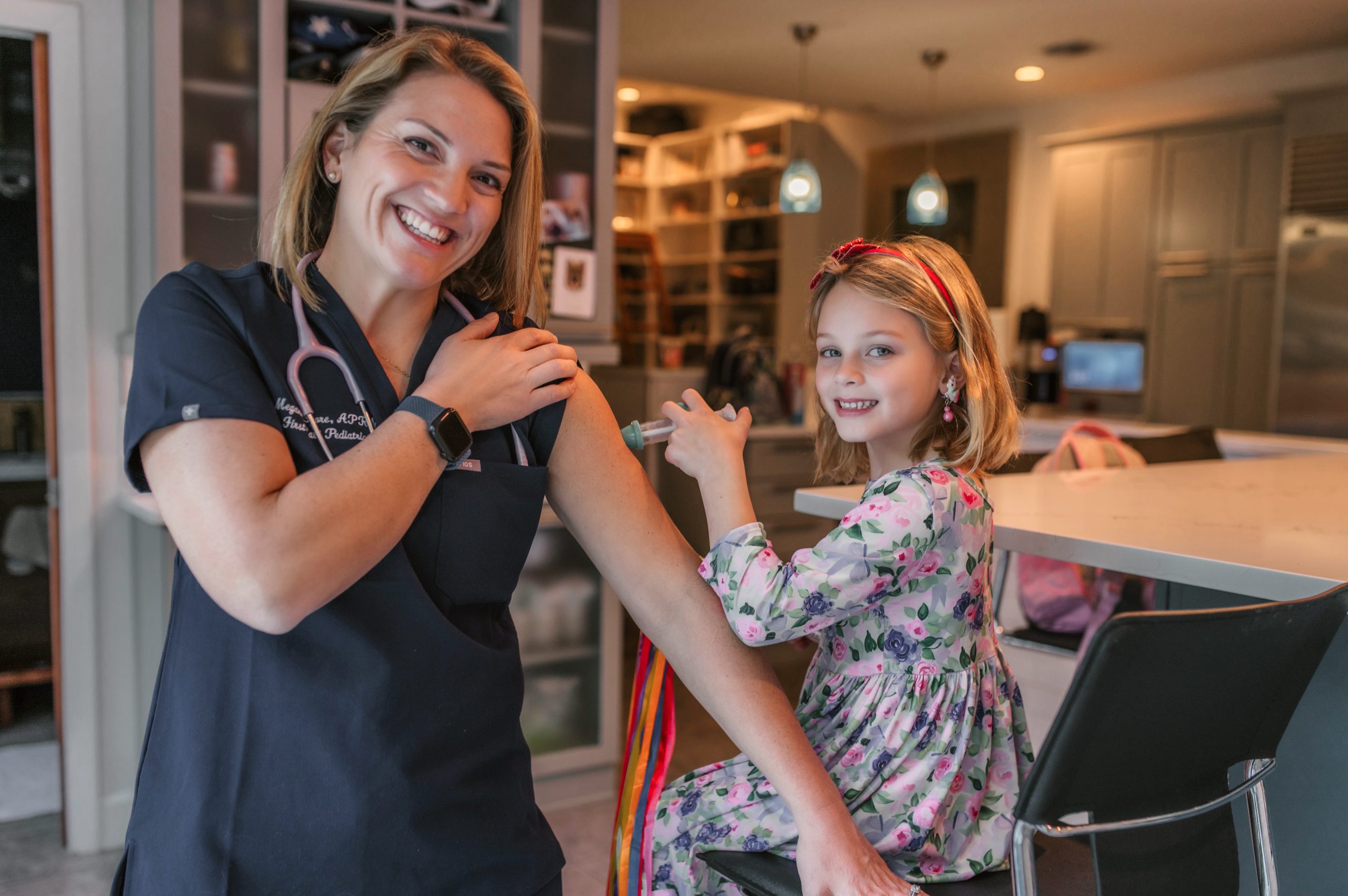 Female pediatric care provider who is wearing First Call Pediatrics scrubs, has her sleeve pulled up, and has a stethoscope around her neck, smiling, standing next to 6-year-old girl, who is smiling, sitting and pretending to give NNP a fake injection with a toy, in-home in San Marco neighborhood in Jacksonville, Fl.