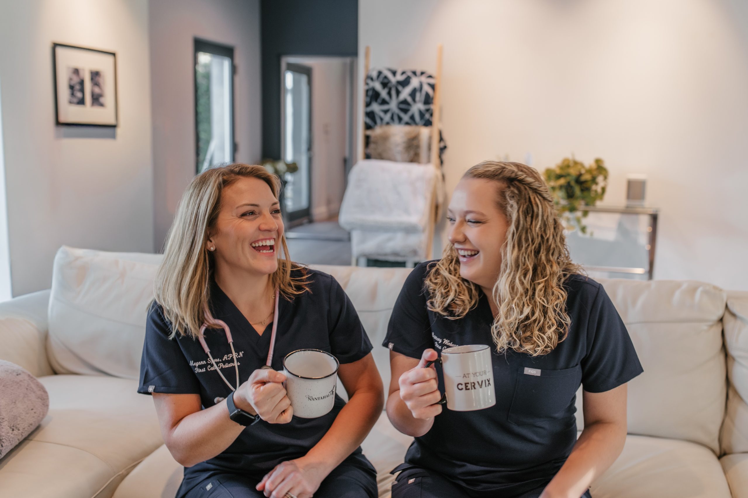 Two female pediatric care providers are wearing First Call Pediatrics scrubs, looking at each other laughing, while holding coffee mugs, with one wearing a stethoscope around her neck, in-home in San Marco neighborhood in Jacksonville, Fl.