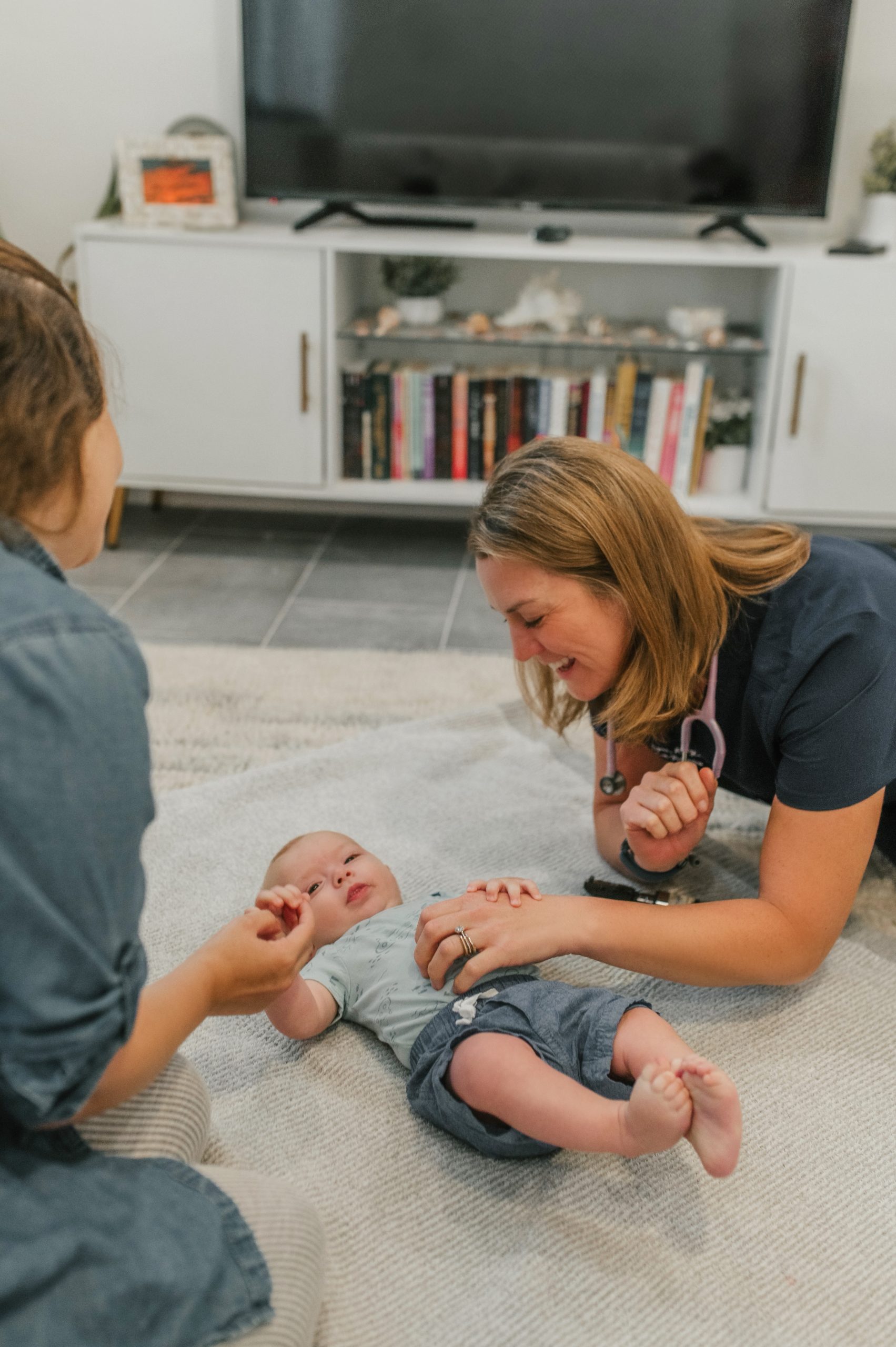 Female pediatric care provider, wearing First Call Pediatrics scrubs, laying on rug, smiling, wearing a stethoscope, tickling an infant boy, while baby holds providers hand, with the mother holds the infant’s other hand, smiling, in-home at Jacksonville, Fl.
