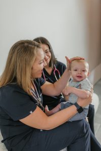 Two female pediatric care providers, wearing First Call Pediatrics scrubs and stethoscopes, sitting next to each other, smiling at baby, while one provider preforms a physical exam on baby’s head with left hand, while the infant, smiling, is being held on the same provider’s lap, in-home in Jacksonville, Fl.