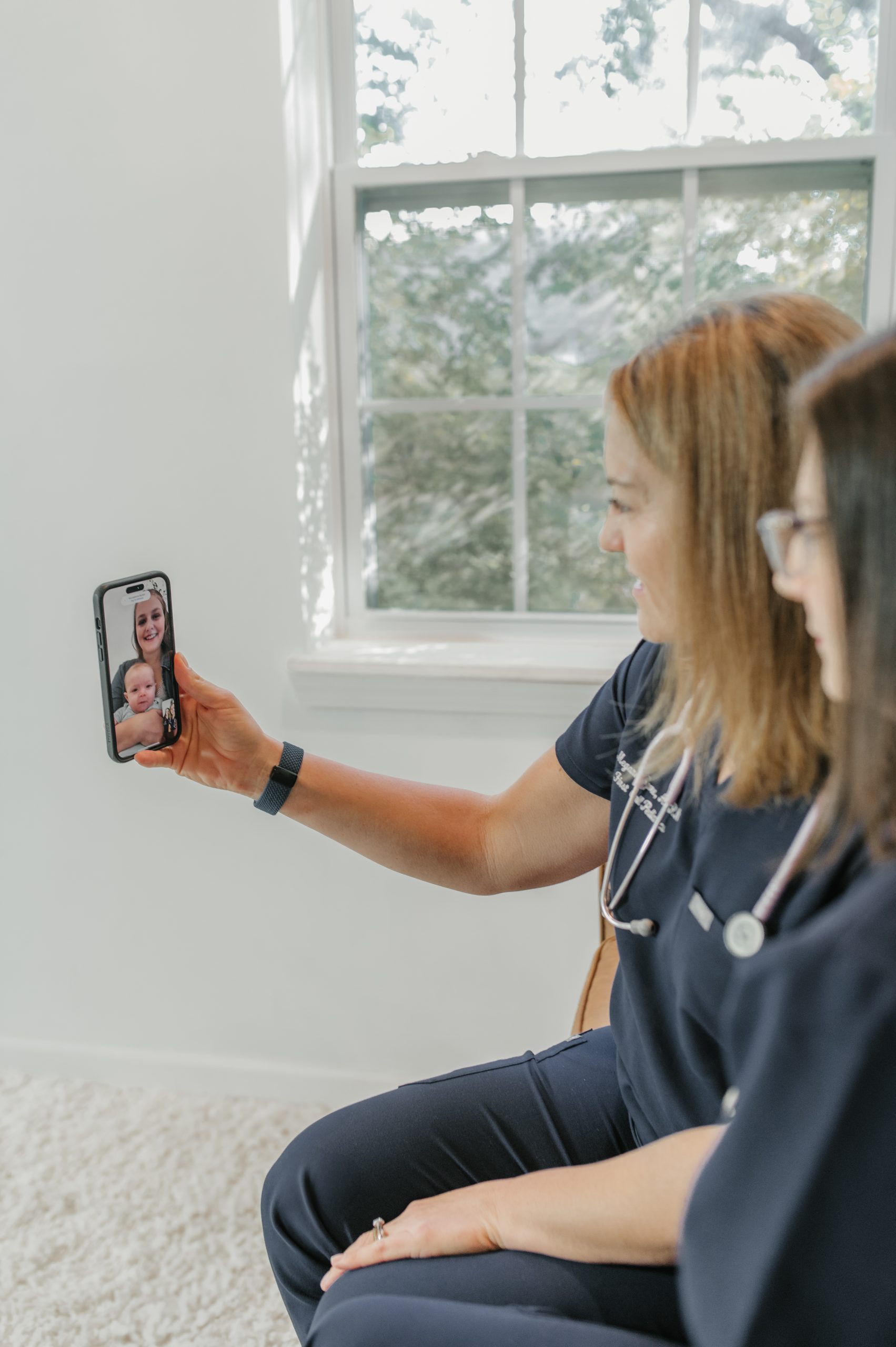 Two pediatric care providers, wearing First Call Pediatrics scrubs and stethoscopes, smiling, look at phone with smiling mother holding her infant on screen, in-home in Jacksonville, Fl.