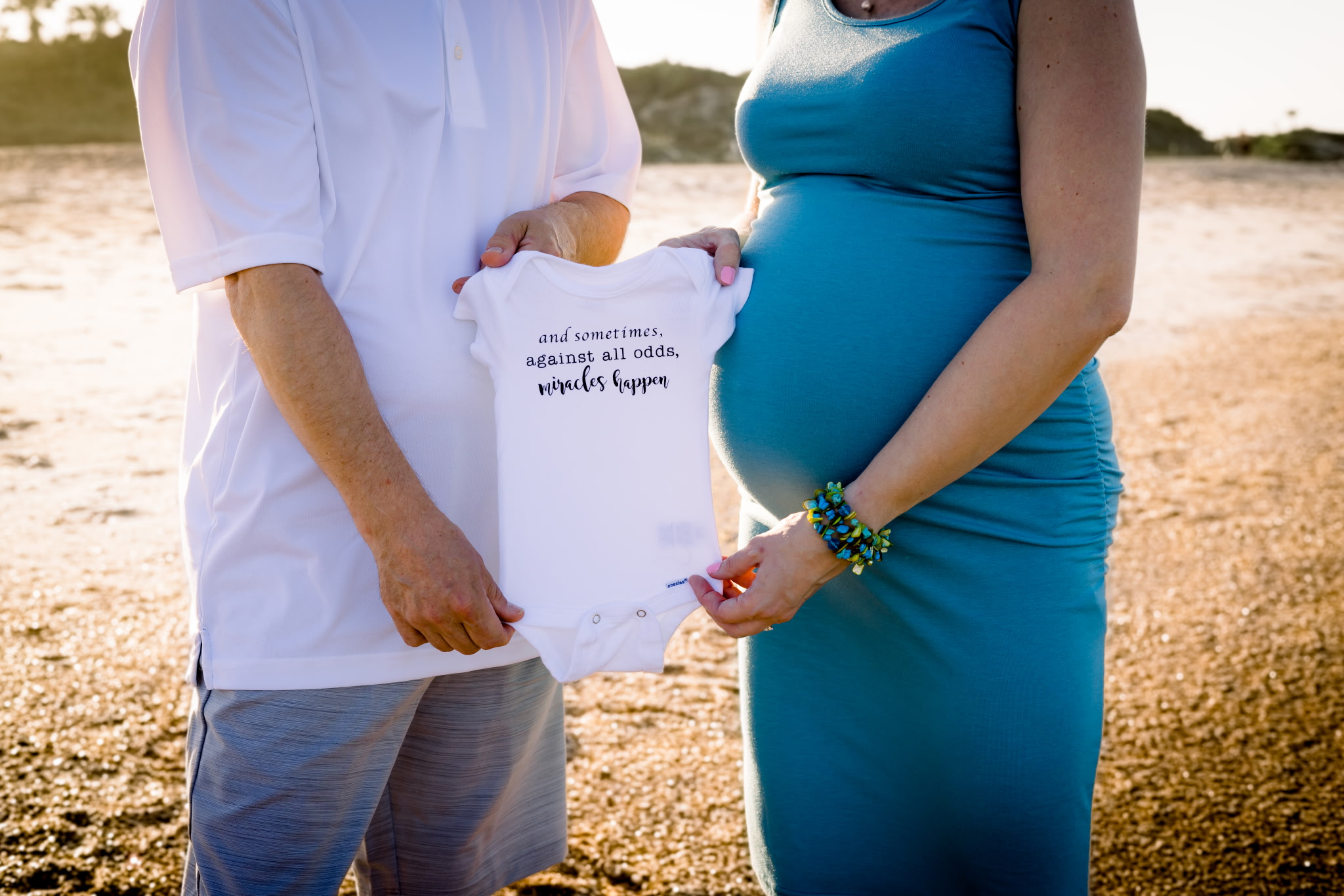 Man in white shirt and blue shorts is standing next to pregnant woman in blue, each holding the side of a onesie together, in Jacksonville Beach, FL