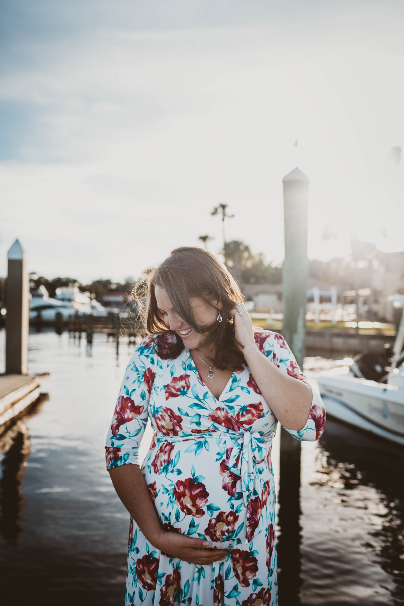 Pregnant woman in a flower-patterned dress, smiles, looking down, with left hand pushing her hair back and her right hand cradling her baby bump, is standing in front of the water, with boats docked in the background, in Riverside, Jacksonville, Fl