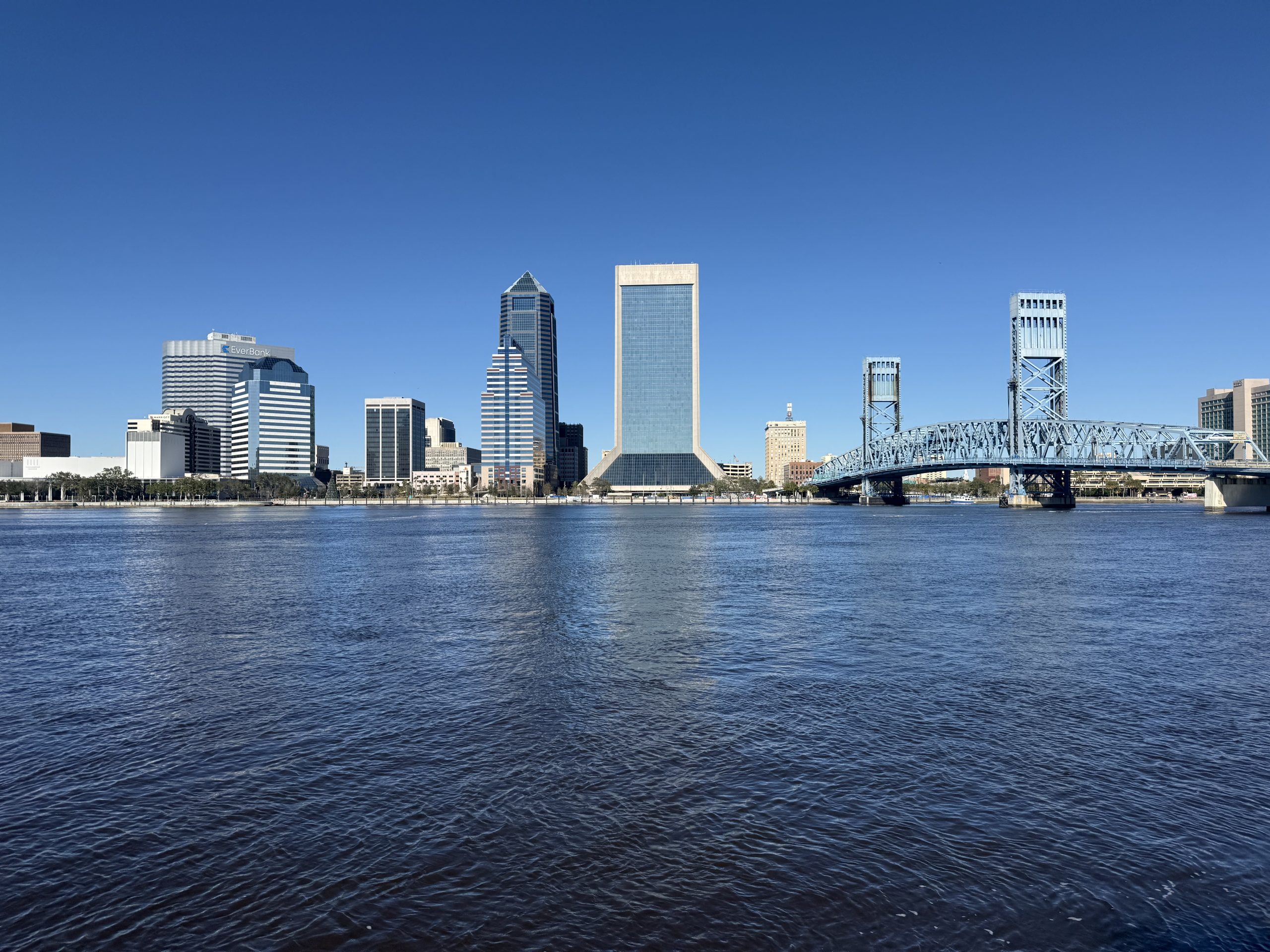 Downtown Jacksonville, FL skyline with several buildings lined up, leading to the Main Street Bridge on the right, with the St. Johns River in the front of the buildings.