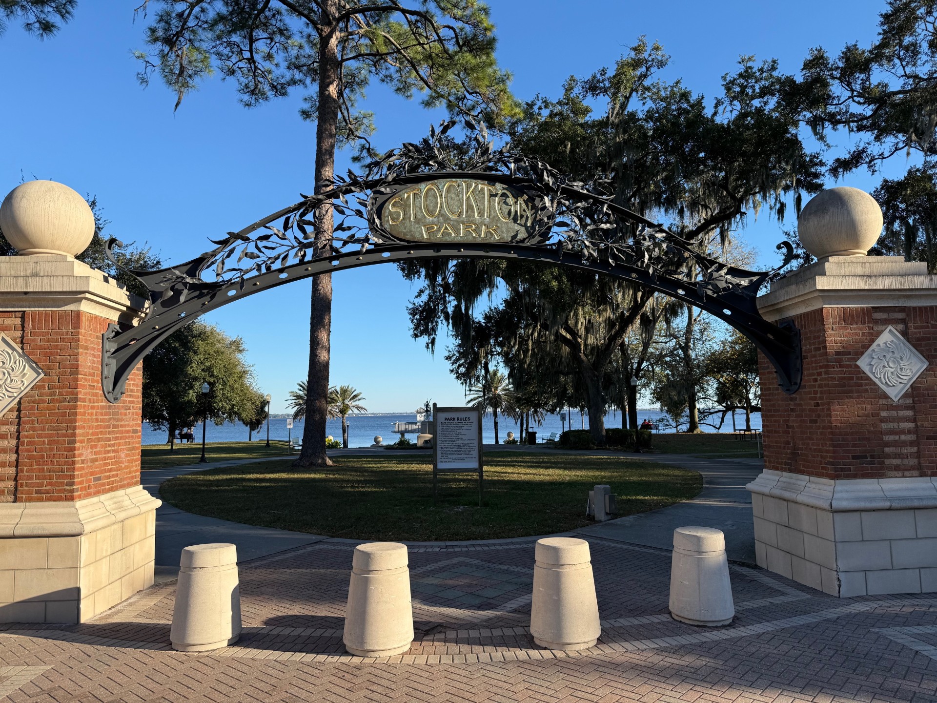 Stockton Park entrance that has a brick pillar on the left and right side, with a metal black arch with leaves crisscrossing through it, with four small stone pillars in front of the walkway and the sign rules with trees surrounding it and the St. Johns River located in the background, located in Ortega, Jacksonville FL.