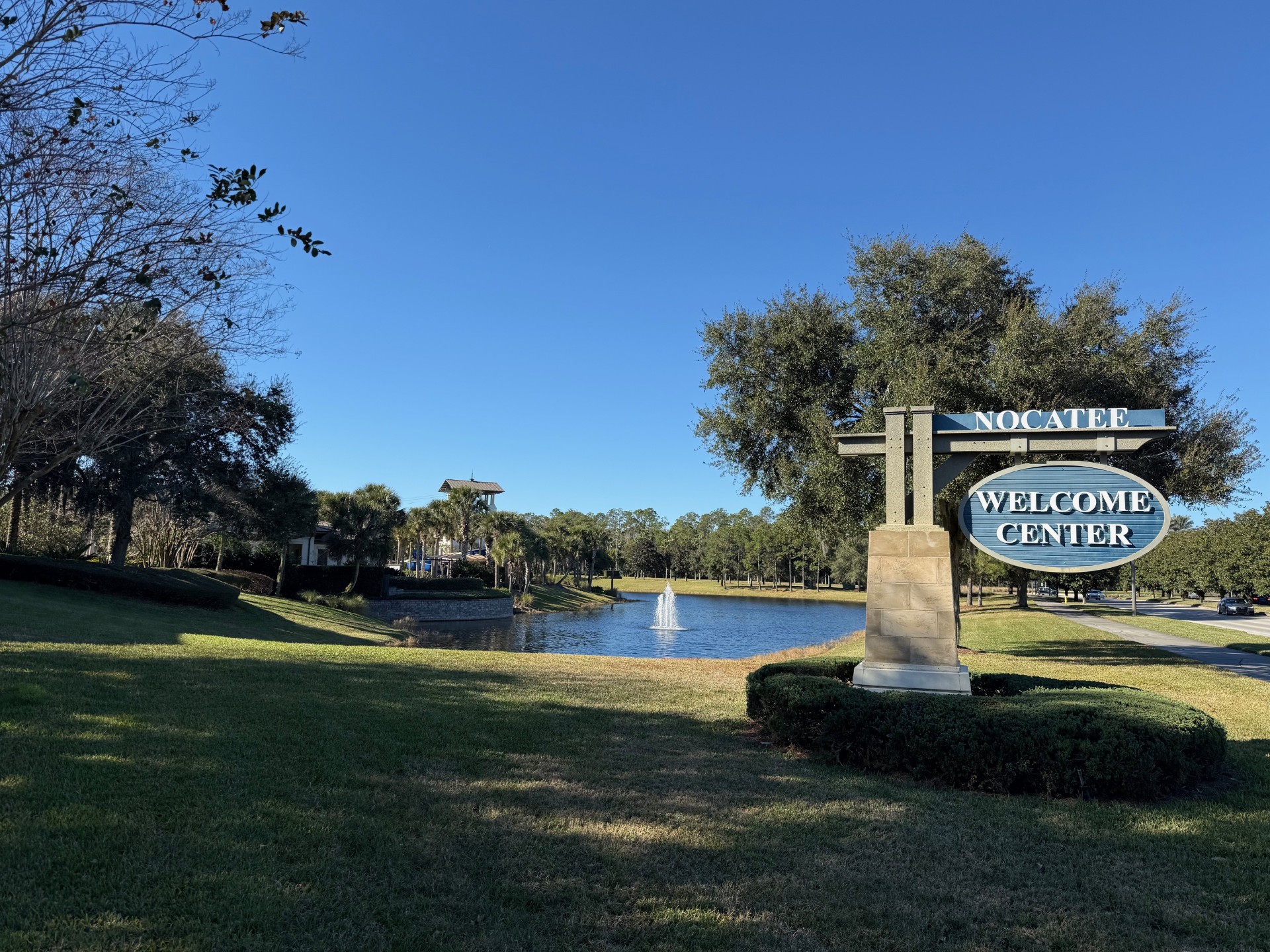 Nocatee Welcome Center Sign on the right with a circle of bushed around it, with a fountain in a pond behind it, and the top of a water slide behind the fountain, surrounded by trees, in Nocatee, FL.