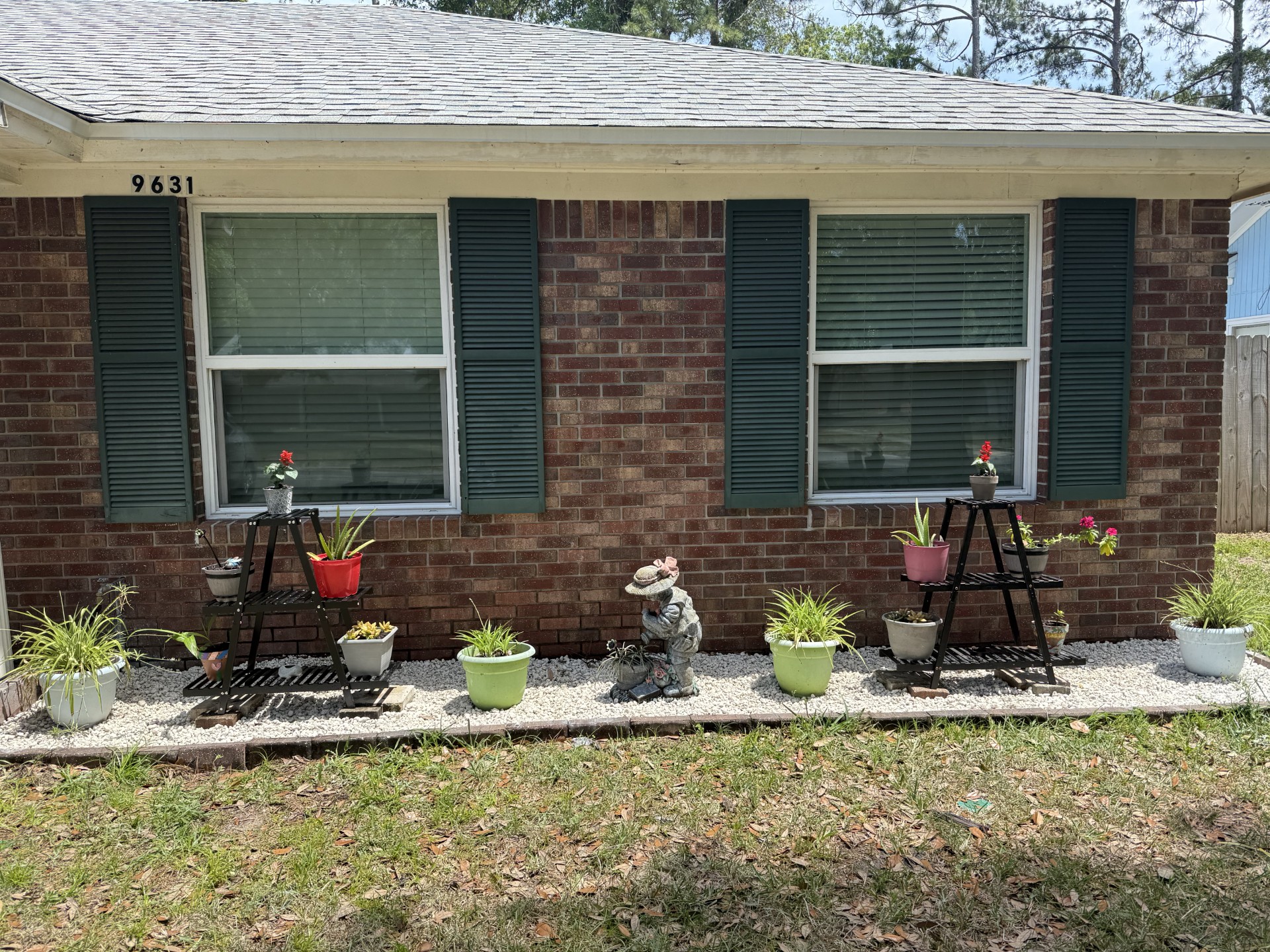 Front of a brick house with two windows with green shutters, a rock garden bed in the front with green plants alternating between two plant stands with flowers on each level with a little girl statue in the middle and grass in front of the garden bed.
