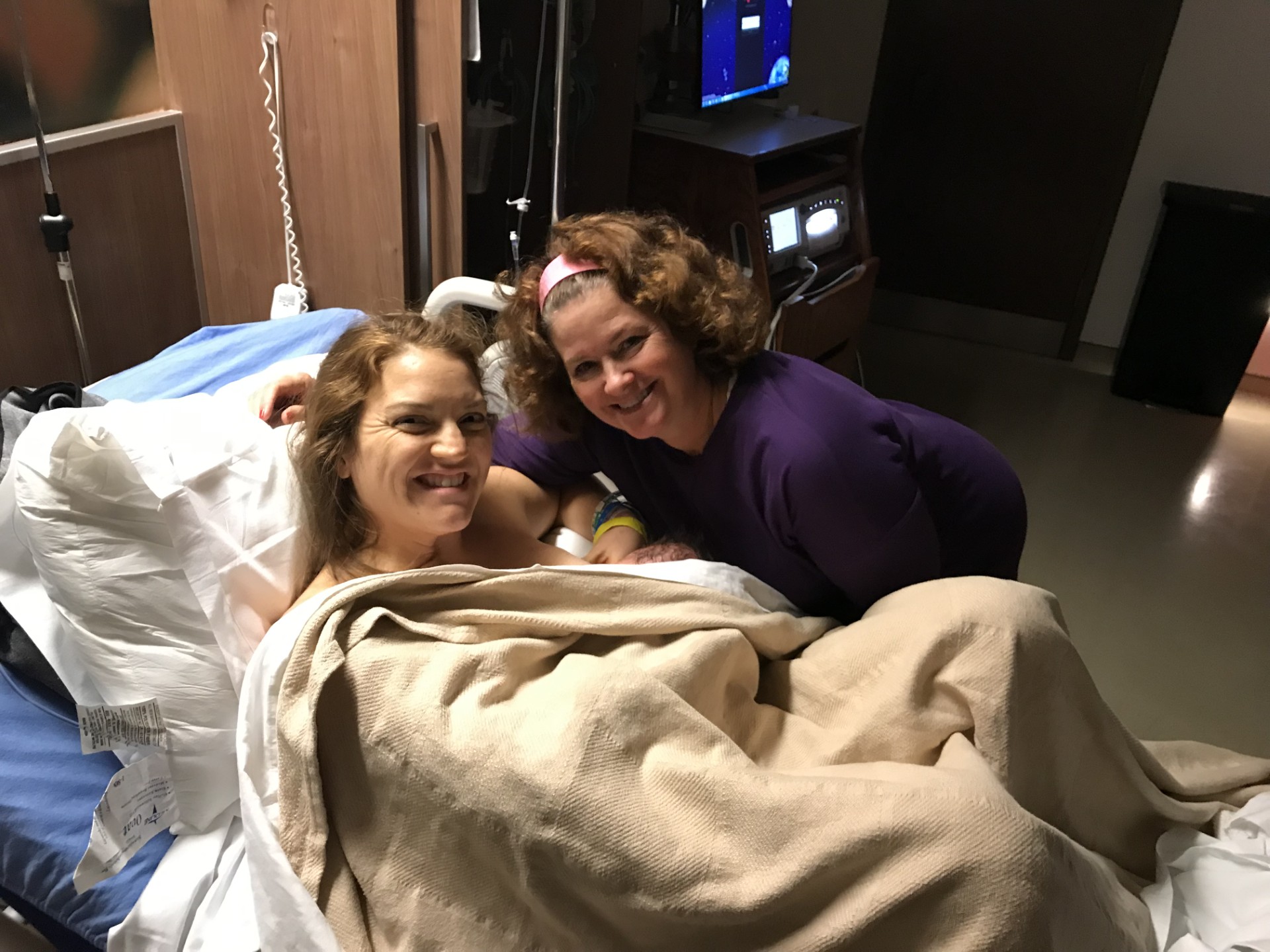 Doula smiling at camera hugging a mother who is holding a baby who is breastfeeding, with a blanket surrounding them both, laying in a hospital bed at UF Health in Jacksonville, FL
