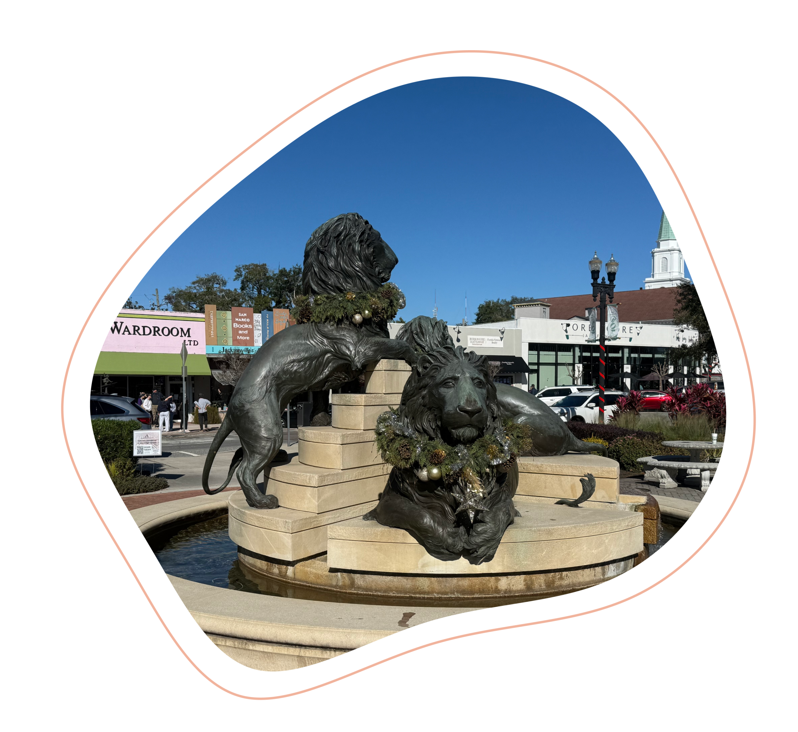 The San Marco statue of three lions positioned in a circle with holiday wreaths on their necks, with one looking straight at the camera, with the fountain water surrounding them in a circle, with bricks surrounding the statue and store fronts in the background, in Jacksonville, FL.