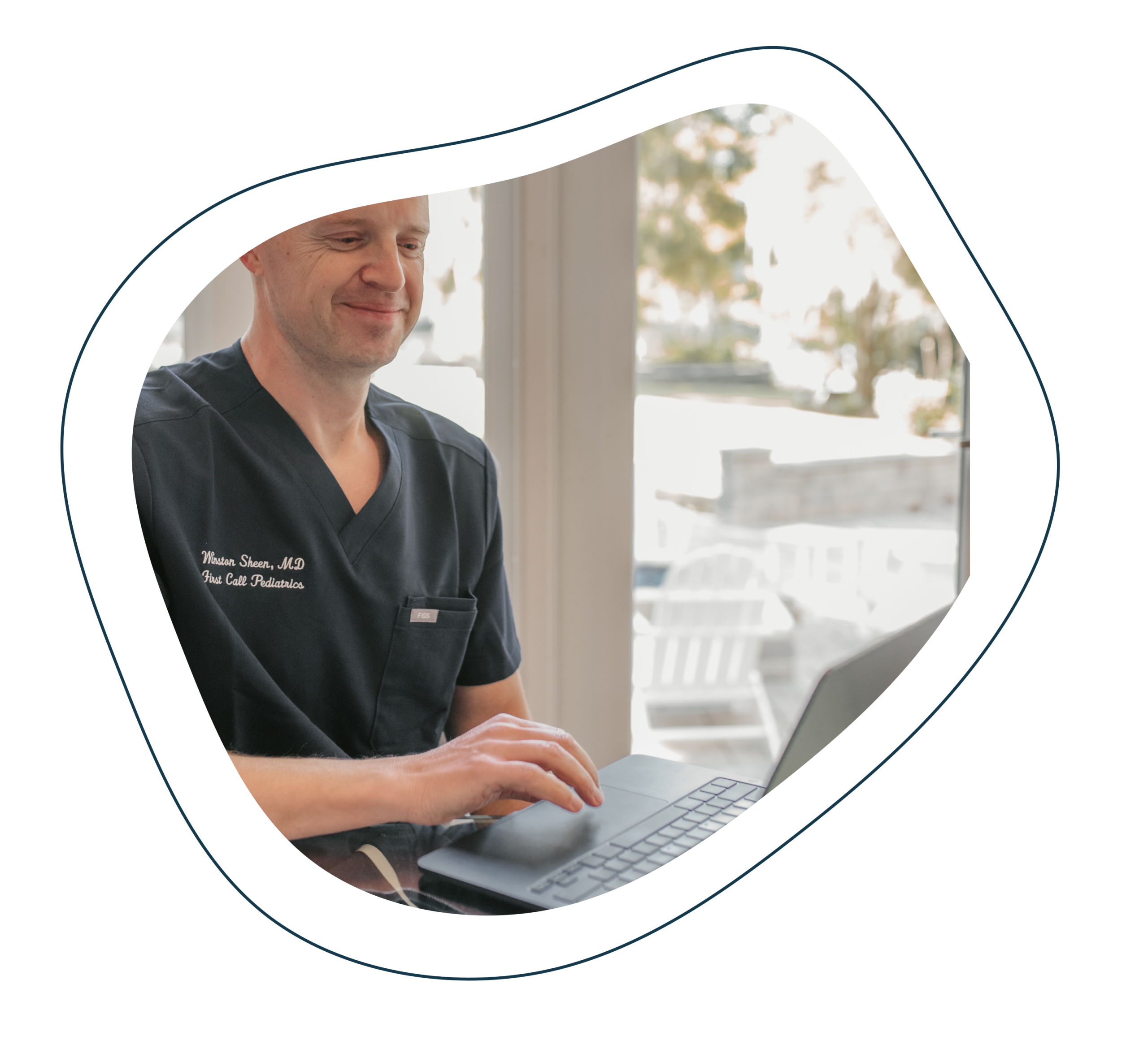 Male Pediatric Doctor is in First Call Pediatric scrubs, smiling, while using laptop in-home in San Marco neighborhood in Jacksonville, Fl.