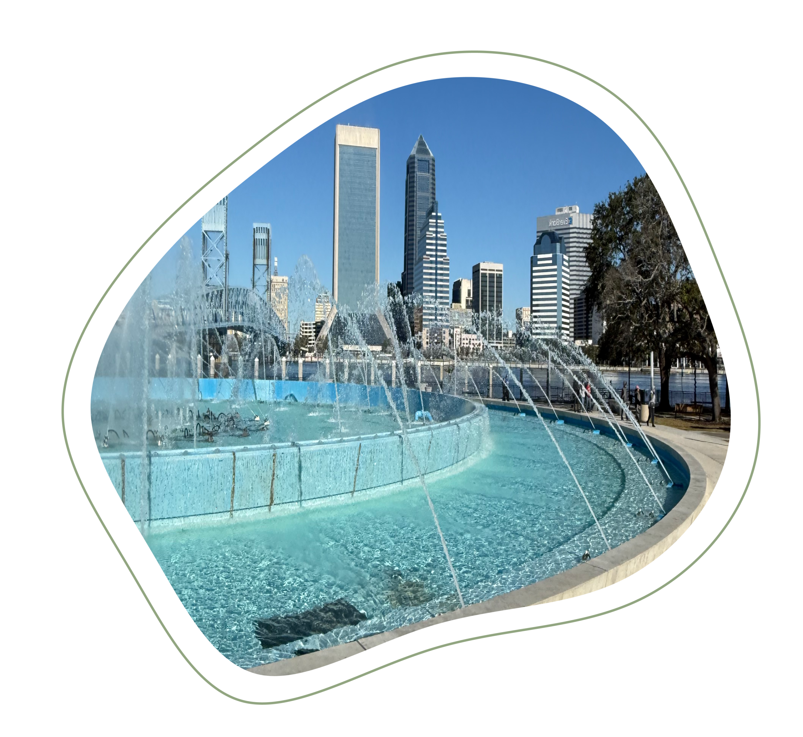 Friendship fountain in downtown Jacksonville, FL is turned on with trees to the left, cement circling the fountain, with the Jacksonville skyline in the background and the Main Street Bridge to the right.