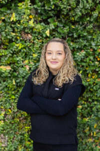 Britteny, Pediatric NP, from the waist up, smiling, with arms crossed and curly blonde hair just past her shoulders, in black First Call Pediatric scrubs and a black long sleeve shirt, with green vines covering the entire background, in San Marco, Jacksonville, FL.