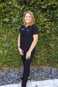 Megann, Neonatal NP, standing on river rocks with right foot and left foot is in front of her on marble tiling, is smiling, with blonde shoulder length, straight hair, in black First Call Pediatrics scrubs, with her left handing resting on left thigh and her right hand resting on right thigh, wearing a watch on right wrist, and green vines covering the entire background, in San Marco, Jacksonville, FL.