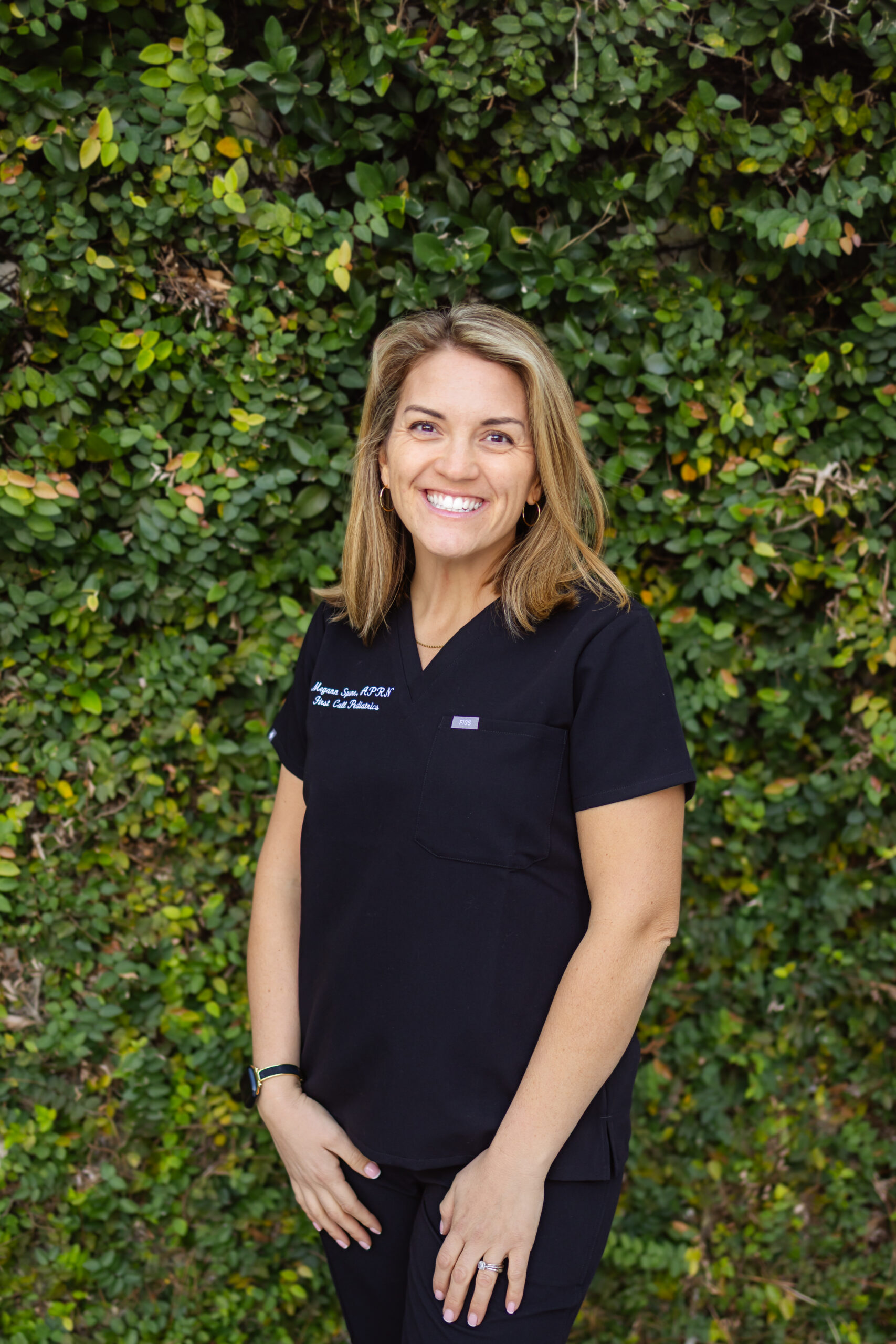 Megann, Neonatal NP, from the waist up, is smiling, with blonde shoulder length, straight hair, in black First Call Pediatrics scrubs, with her left handing resting on left thigh and her right hand resting on right thigh, wearing a watch on right wrist and ring on her left fourth finger, with green vines covering the entire background, in San Marco, Jacksonville, FL.