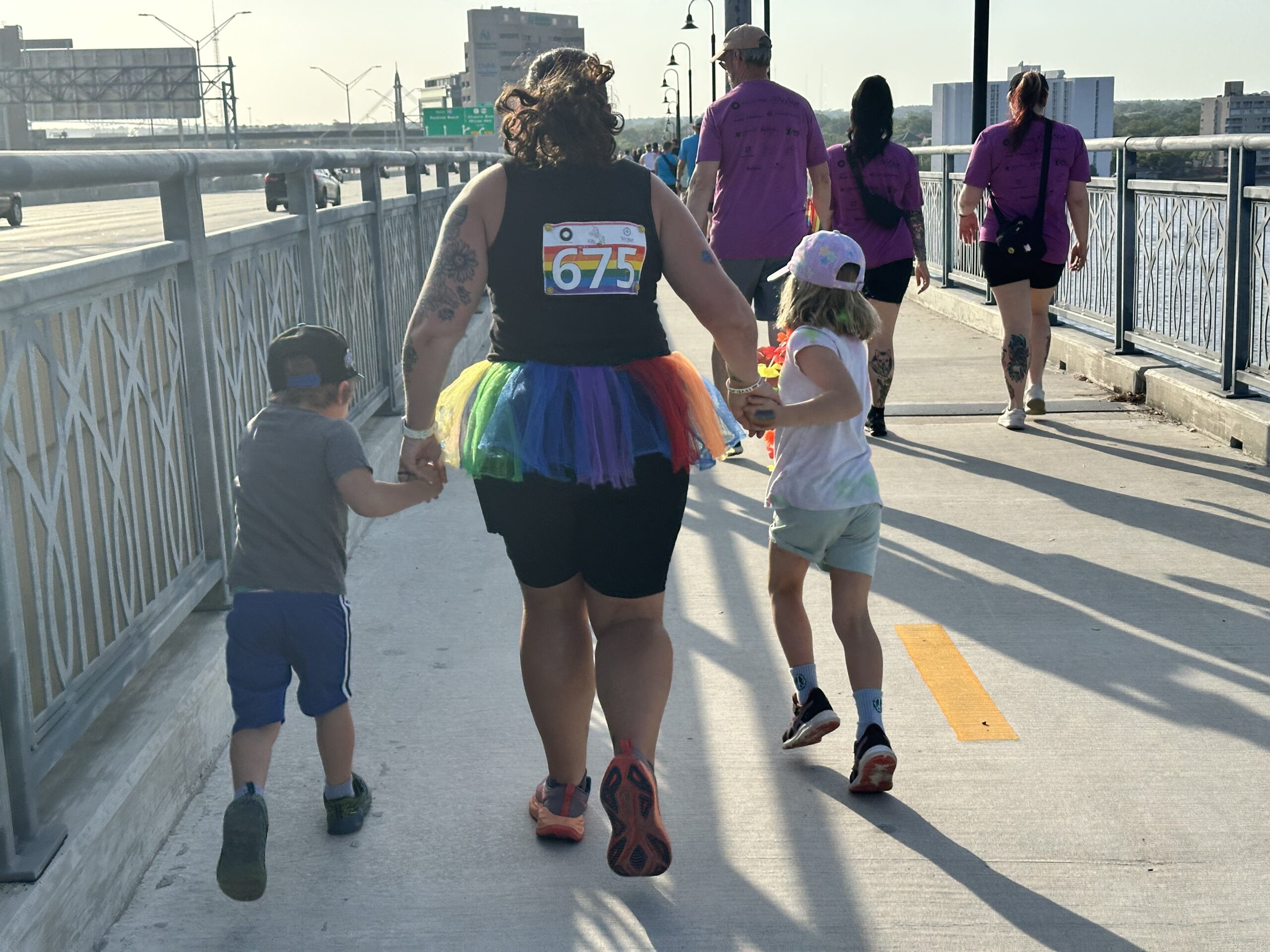 A pediatric aged boy in blue shorts, grey shorts, and a black hat, running while holding a woman in a black shirt and shorts, with a rainbow tutu and orange sneakers, with the other hand holding a pediatric aged girl in a white hat, white shirt, light blue shorts, and black sneakers, all running together over the walking path on I95, in Riverside, Jacksonville, FL