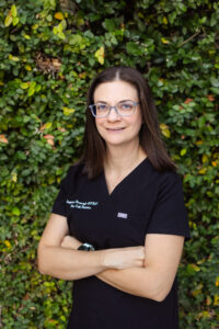 Steph, Neonatal NP, from the waist up, arms crossed, wearing eyeglasses, smiling, with brown shoulder length, straight hair, in black First Call Pediatrics scrubs, wearing a watch on left wrist, with green vines covering the entire background, in San Marco, Jacksonville, FL.