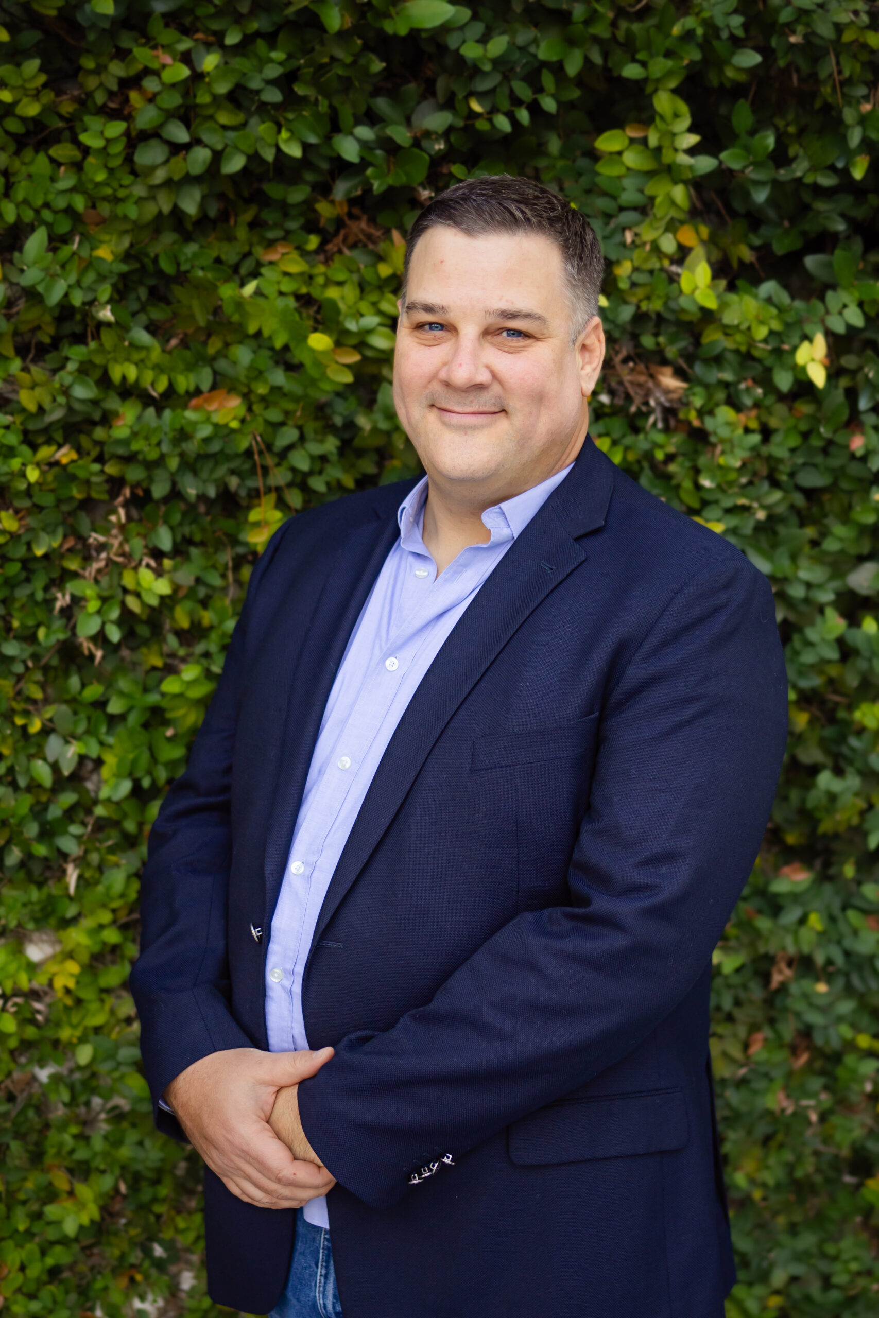 Kyle, from the waist up, hands crossed at hip level, smiling, with brown short hair, in a light blue shirt, with a suit jacket, with green vines covering the entire background, in San Marco, Jacksonville, FL.