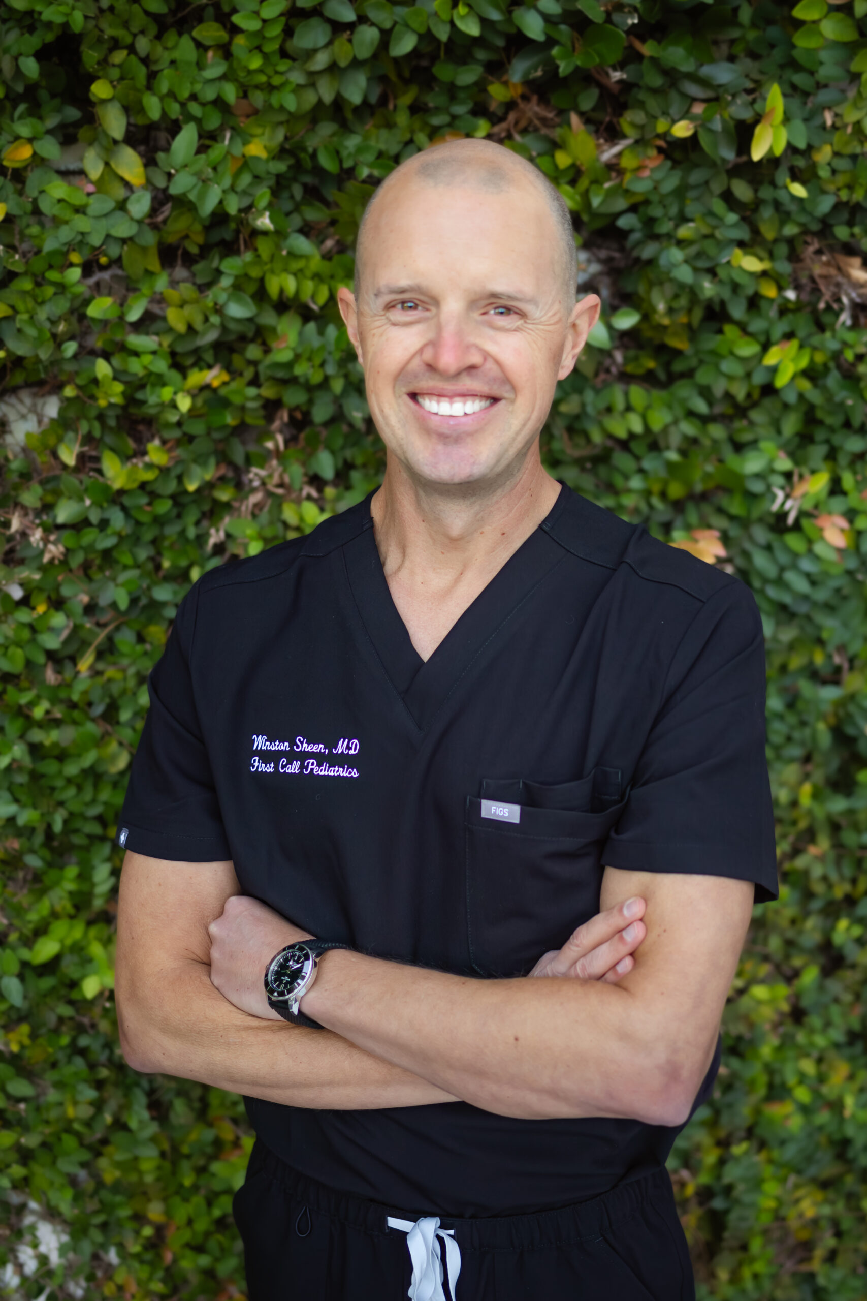Winston, MD, from the waist up, arms crossed, smiling, in black First Call Pediatrics scrubs, wearing a watch on left wrist, with green vines covering the entire background, in San Marco, Jacksonville, FL.