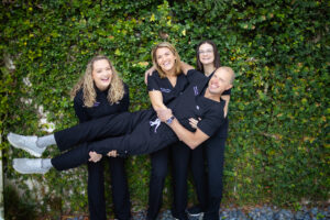 First Call Pediatrics medical team of four in black team scrubs, with Winston, MD wearing a watch on left wrist, smiling, with his arm around middle person’s neck, being held by Britteny, Pediatric NP, holding his legs, smiling with curly blonde hair past her shoulders, and wearing a black long sleeve shirt under her scrubs, Megann Neonatal NP in the middle holding his stomach, with shoulder length blonde hair, smiling, next to next to Steph, Neonatal NP, holding his back, smiling, with brown shoulder length, straight hair, with green vines covering the entire background, in San Marco, Jacksonville, FL.
