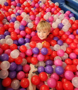 Pediatric aged boy with blonde hair smiling in a ball pit, completely covered except for hear, with balls that are red, purple, white, and pink.