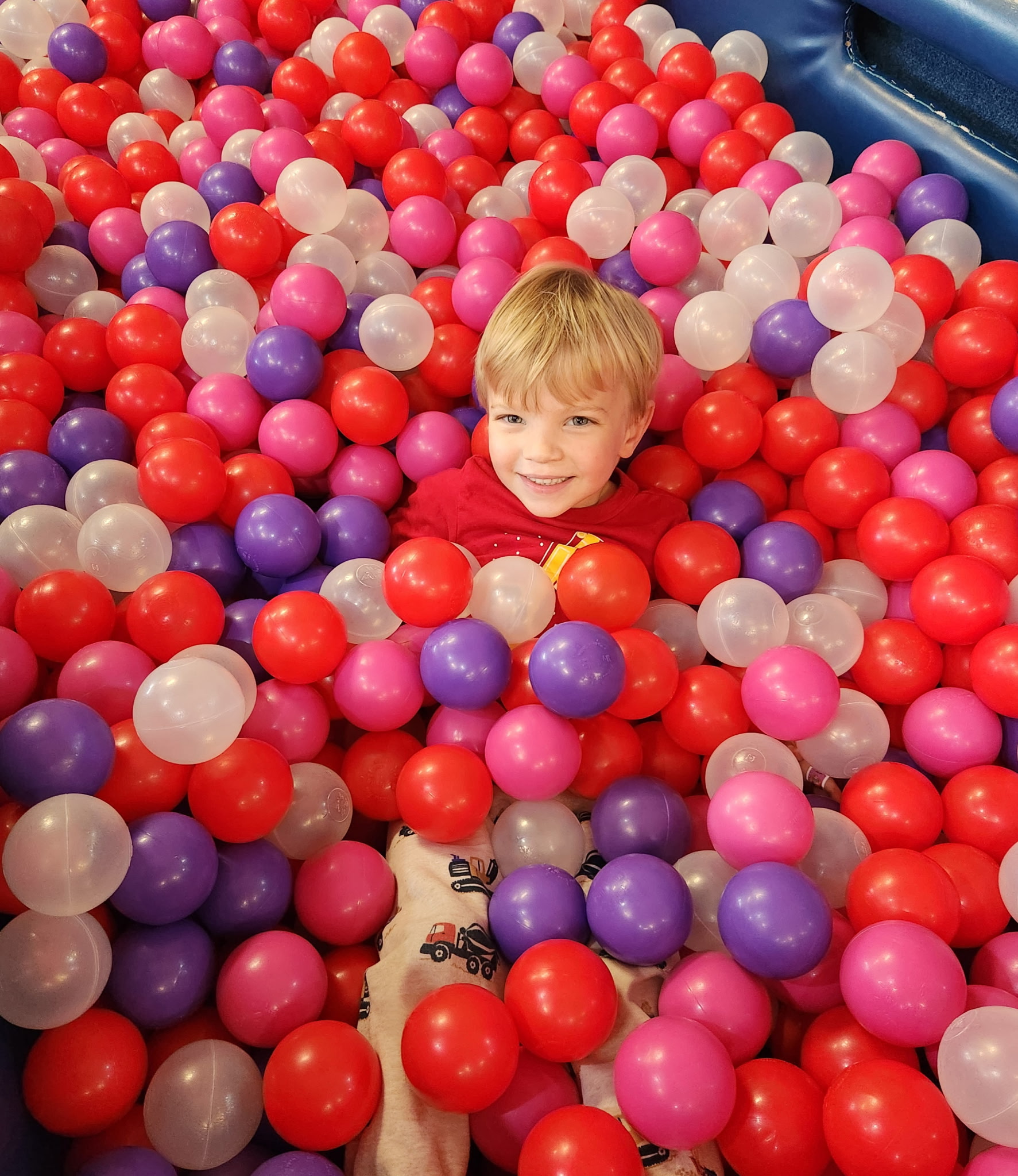 Pediatric aged boy with blonde hair smiling in a ball pit, completely covered except for hear, with balls that are red, purple, white, and pink.