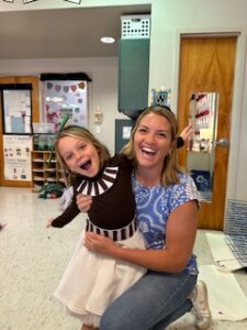 Woman smiling on the right, wearing a blue and white patterned shirt and jeans, holding a pediatric girl wearing a brown shirt and white skirt, smiling, in a school classroom, in Riverside, Jacksonville, FL.
