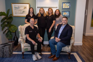 First Call Pediatrics team of six, with Winston, MD on the far left, wearing a watch on left wrist, smiling, sitting on an off-white patterned fabric chair, with Steph, Neonatal NP, standing behind his chair with her arm on the back of the chair, smiling, with brown shoulder length, straight hair, next to Britteny, Pediatric NP, standing, smiling with curly blonde hair past her shoulders, and wearing a black long sleeve shirt under her scrubs with her arm around Steph and Megann, with Megann Neonatal NP standing in between the chairs, smiling, with shoulder length blonde hair, with Kat next to her, hand on the back of the chair, in a light purple sweater, smiling, with Kyle sitting on an off-white patterned fabric chair, hands intertwined on lap, smiling, with a blue rug and wood flooring under the chairs, art work on the walls, and a plant on the left of the chairs, in San Marco, Jacksonville, FL.