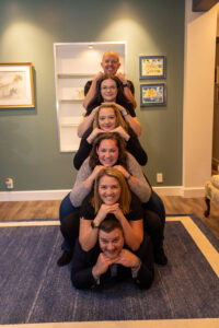 First Call Pediatric team is stacked on upon each other, with hands under chins, with Kyle on the floor, with Megann above him, Kat above her, Britteny above her, Steph above her, and Winston at the top, all smiling, with a blue wall with pictures and an open bookcase in the background in San Marco, Jacksonville, FL.
