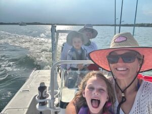 A boat in the water with Megann, neonatal NP, smiling, wearing a sun hat and sunglasses, with a white shirt, holding a pediatric girl with her mouth open, wearing a live vest, with a man behind them driving, wearing a sun hat and sun glasses and white shirt, holding a pediatric boy who is wearing a hat and smiling.