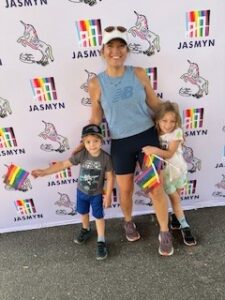 Megann, Neonatal NP standing, smiling, wearing a blue tank top and black short leggings, with arms around pediatric boy on left side, wearing blue shorts and gray graphic t-shirt, and girl on right side, in green shorts and yellow shirt, both holding pride flags, with Jasmyn backdrop behind them in San Marco, FL.