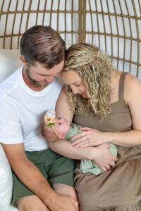 • Man, wearing a white shirt and green shorts, next to Woman in brown dress, together holding newborn on white pillows in an egg chair in Jacksonville, FL