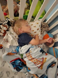 Pediatric boy sleeping in crib with a pillow under head, wearing a blue shirt and a blanket over lap in Jacksonville Beach, FL