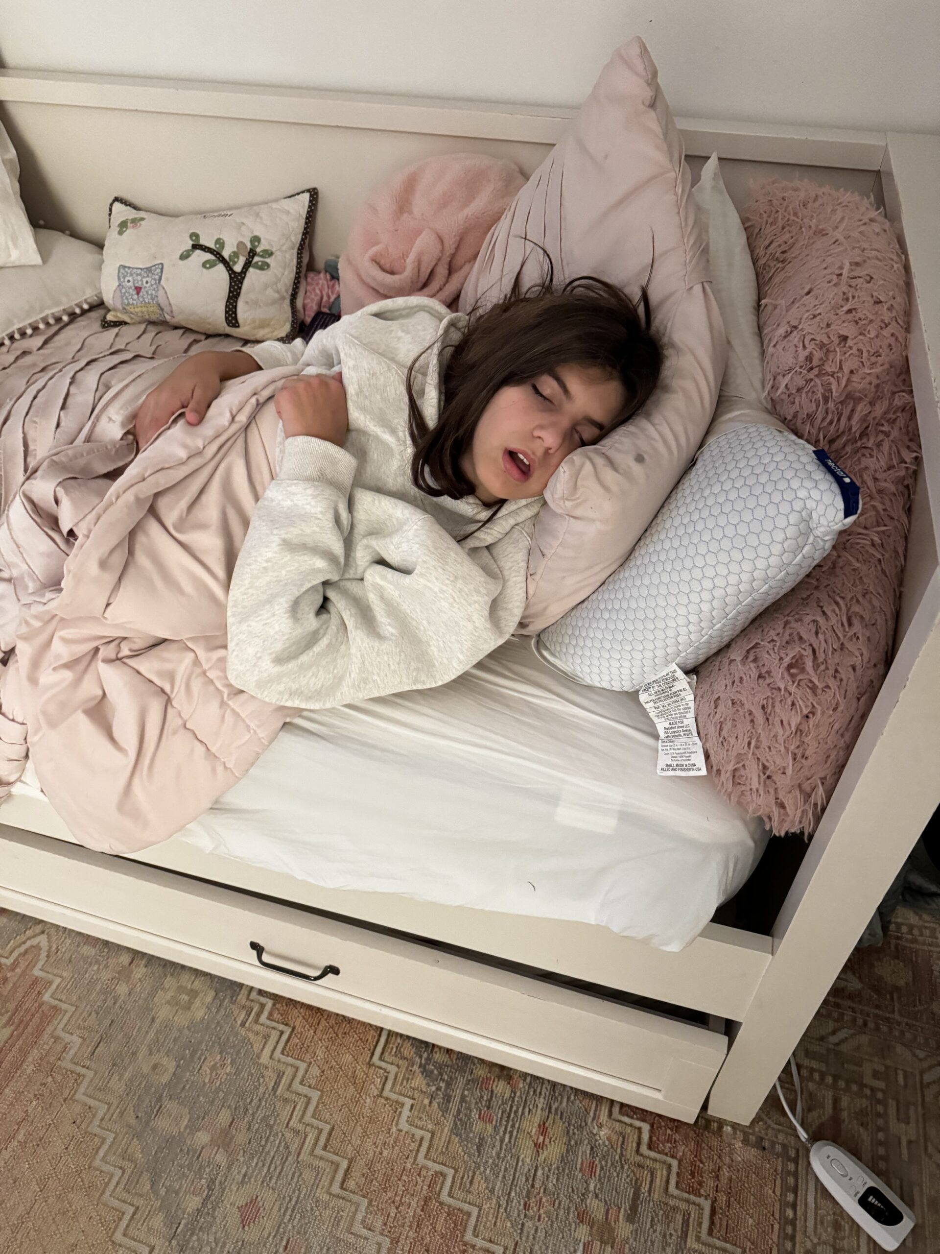 Pediatric girl sleeping in bed with pink pillows stacked behind her, a pink blanket, in a white hoodie, in Nocatee FL