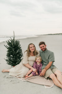 Britteny, pediatric NP, on the left, wearing a white dress, with a toddler smiling in a pink dress, and man in green shirt and white pants on the right, wit a christmas tree to the left and the beach in the background, at Jacksonville Beach, FL