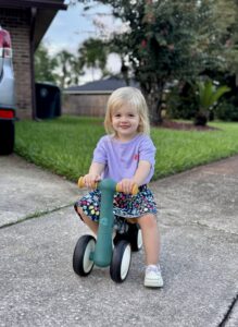 Pediatric aged girl riding a green tricycle with a purple shirt, flowered skirt, and white sneakers, in Jacksonville, FL