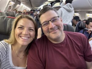 Megann, Neonatal NP on the right, smiling, with blonde straight hair, with Kyle, smiling, wearing a red shirt, on an airplane, and people in the background, in Jacksonville, FL.