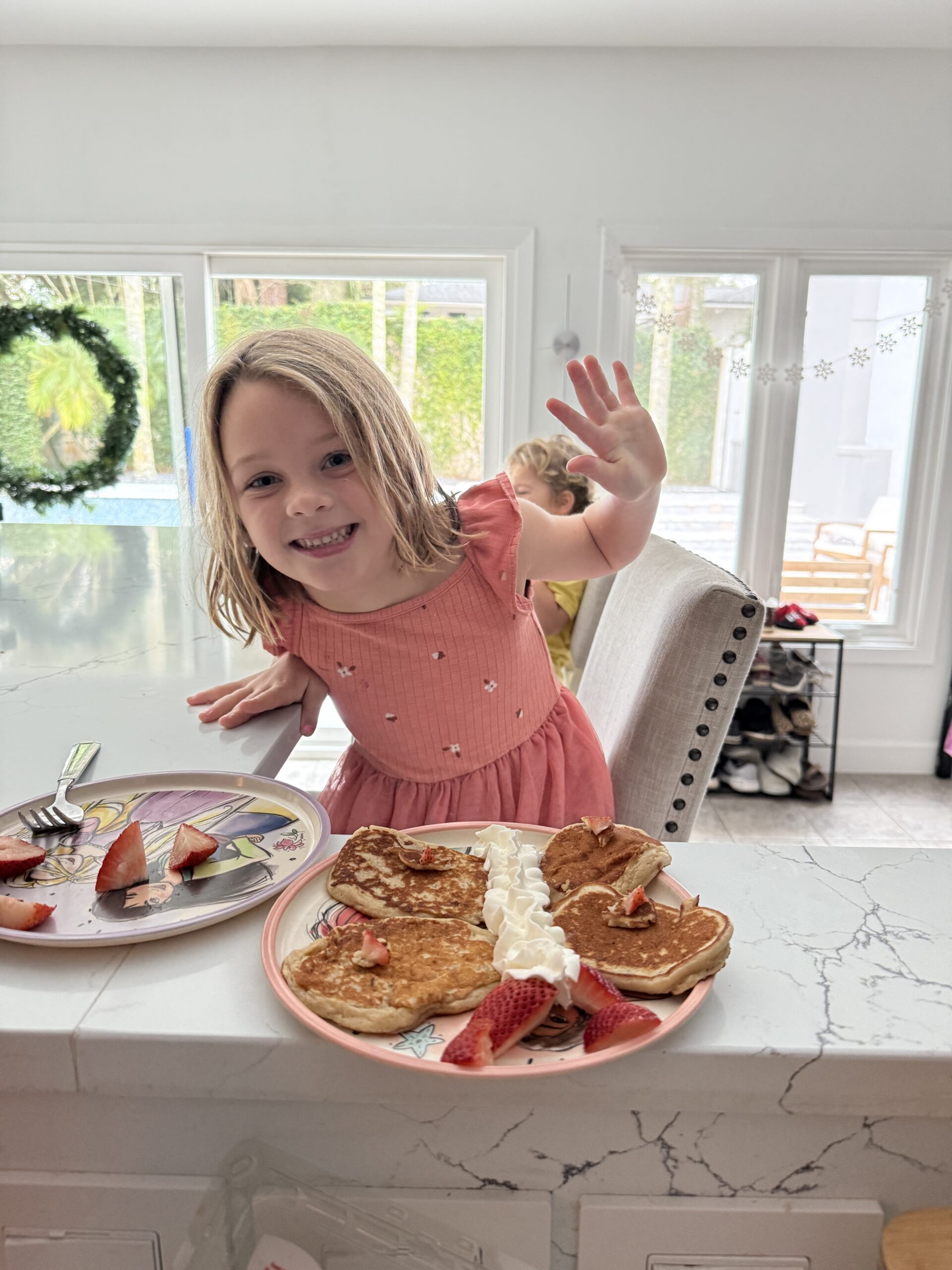 Pediatric aged girl, smiling, in pink dress with pancakes as wings and strawberries as the antenna to make a butterfly, with large sliding glass doors in the background, in San Marco, Jacksonville, FL