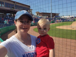 Woman, smiling, wearing a blue baseball hat and white shirt, holding a pediatric aged child, wearing a red shirt with blonde hair, at baseball stadium in Jacksonville, FL