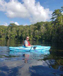 Woman in a blue kayak and wearing a red life vest and grey shirt, kayaking in water with trees behind her