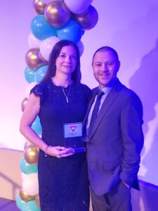 Woman, in a dark blue dress and necklace, smiling, holding an award standing next to a man in a grey suit and blue tie, with a balloon arch behind them, in Jacksonville, FL