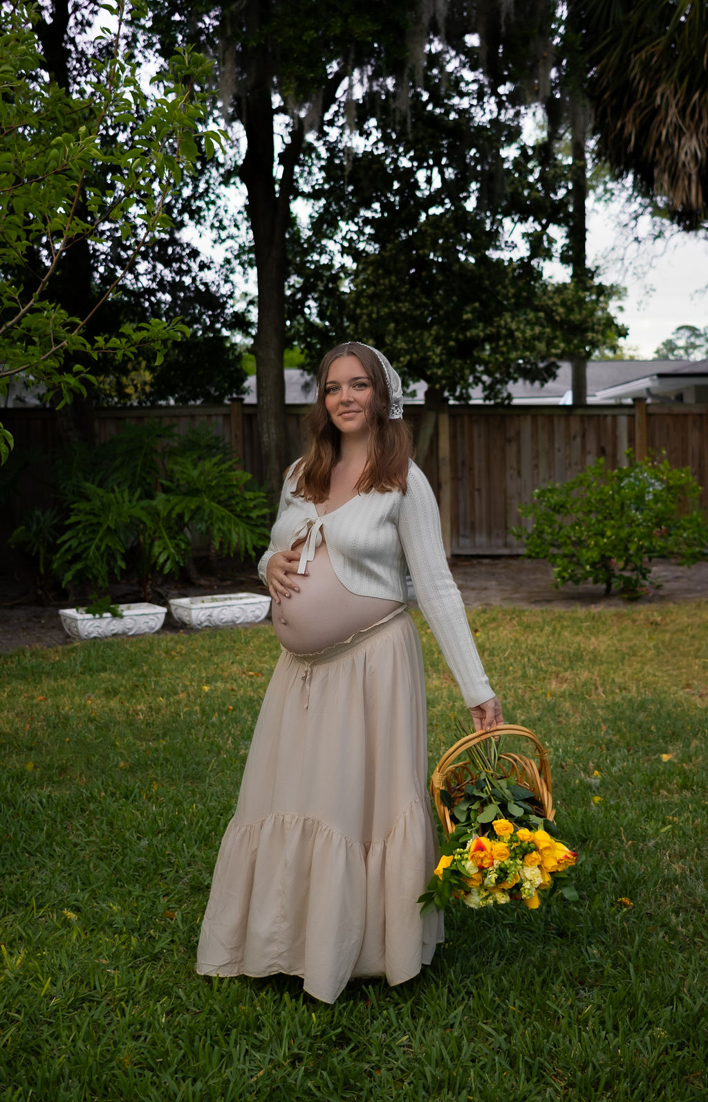 Pregnant woman in white sweater with hand on belly, tan skirt, and right hand holding a basket with sunflowers standing in grass.