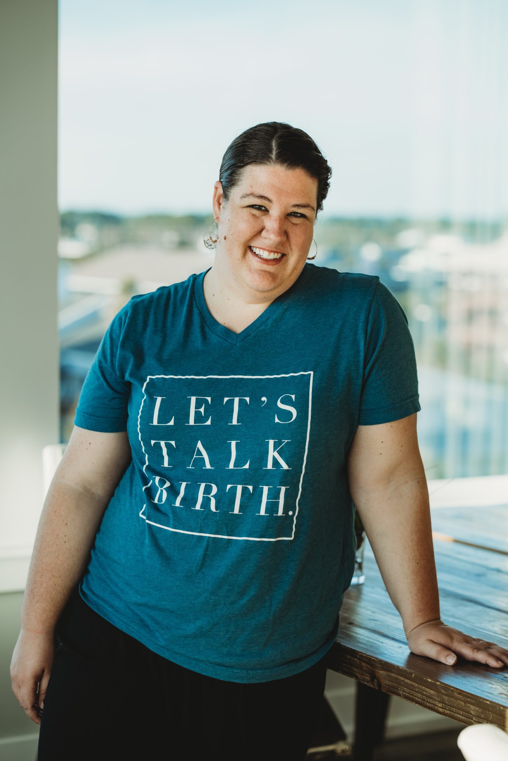 Woman smiling at camera, in a blue shirt that says let's talk birth with black pants and dark brown hair, with a window and buildings in the background.